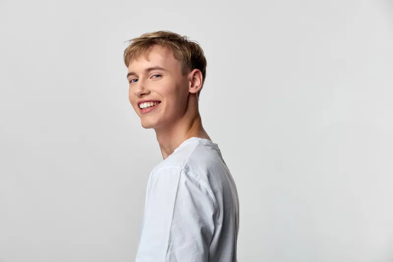 Smiling young man in a white casual shirt stands against a plain background, radiating friendliness and confidence for lifestyle, fashion, and corporate branding projects in a clean studio setting