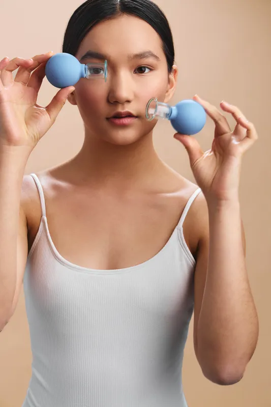 Asian woman holds small dumbbells near face in a focused home workout moment. She smiles slightly, while maintaining balance and alignment, creating a calm, energetic fitness scene.