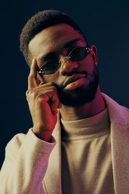 Stylish young black man in sunglasses with thoughtful expression, wearing a light colored sweater and blazer, against a dark blue background