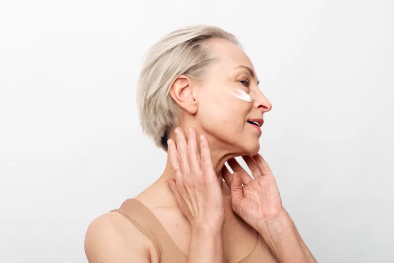 Mature woman with short grey hair applies moisturizing cream on face and neck on white background. Concept of skincare, aging skin care, beauty routine, and natural self-care for older adults.