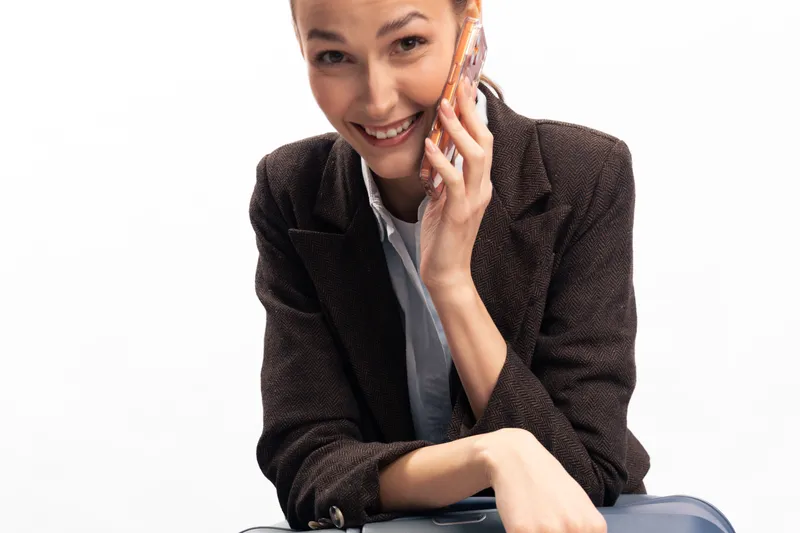 Smiling woman in brown jacket talking on smartphone, sitting with suitcase on white background. Concept of travel, communication, business trip, and positive mood.