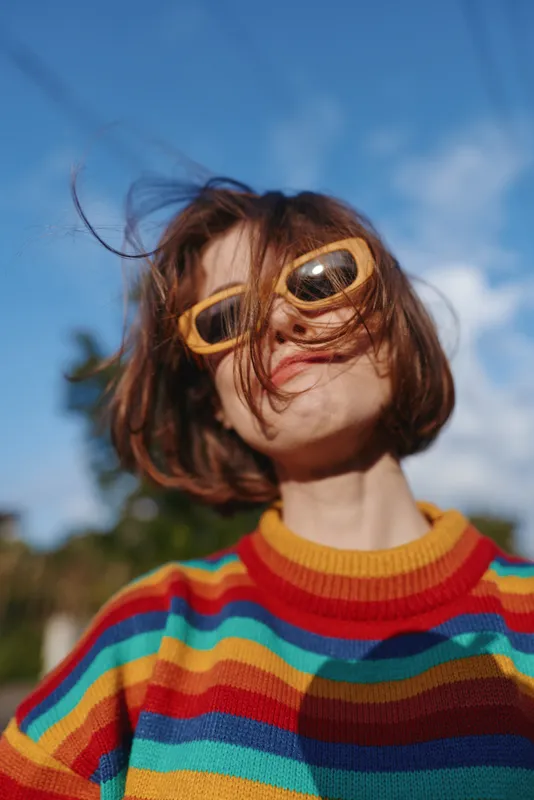 Young woman in rainbow sweater and yellow sunglasses, smiling with windblown hair in outdoor portrait against tropical blue sky; playful travel lifestyle and casual fashion.