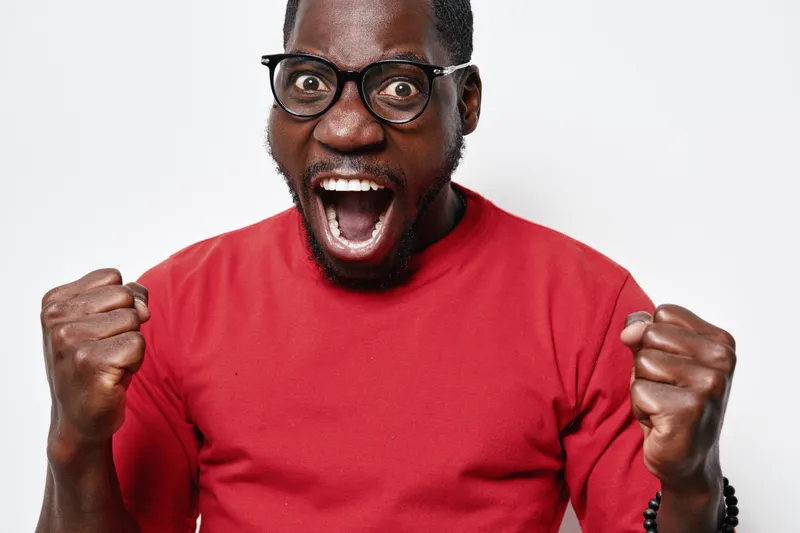 African American man with dark skin and glasses wearing red shirt expressing excitement with clenched fists and open mouth on white background. Emotion, joy and celebration.