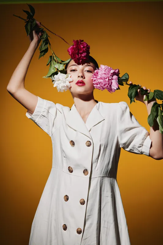 Young woman in white dress holds flowering branch, wearing red lipstick and creative flower headpiece. Vibrant yellow background complements beauty fashion concept.