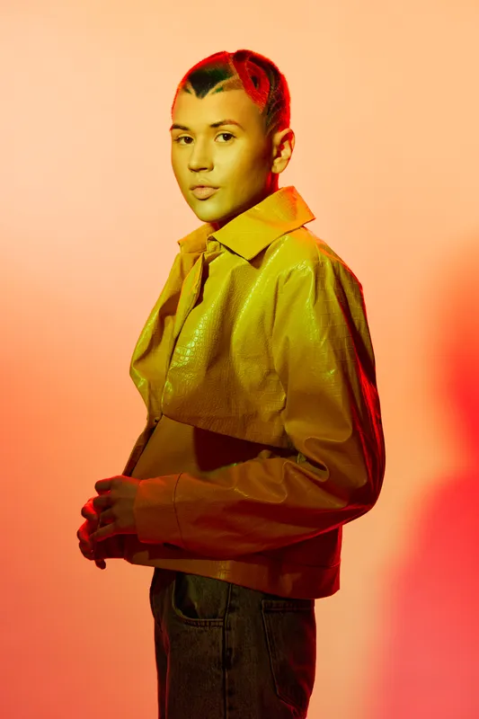stylish woman with braided hair wearing yellow jacket stands against warm orange background, showcasing modern fashion and confident pose in studio lighting
