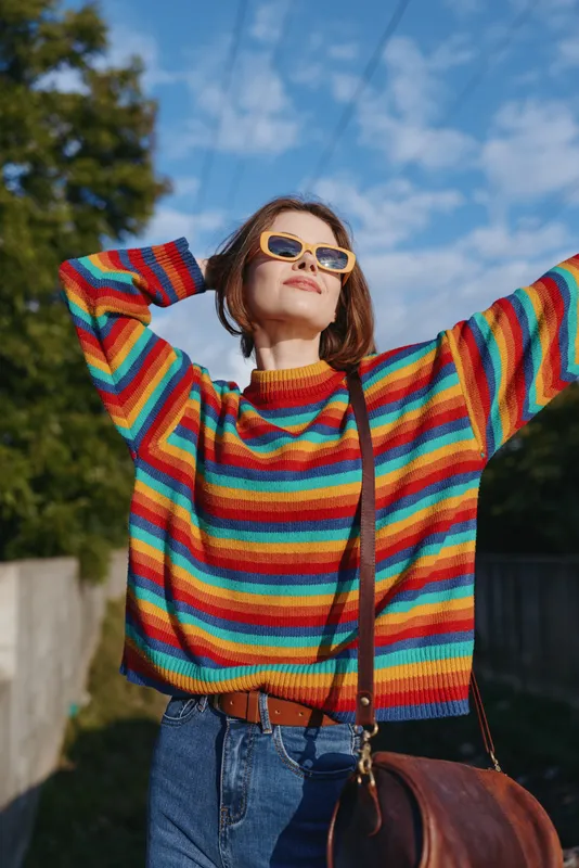 woman in rainbow sweater and sunglasses, jeans and shoulder bag outdoors, smiling and raising arms with confident relaxed pose under blue sky, casual travel lifestyle, 25-year-old female with bob