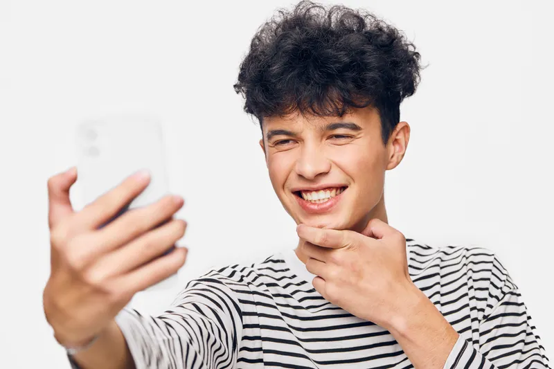 young man taking selfie with smartphone, smiling and wearing striped shirt against white background, casual and cheerful mood captures modern youth lifestyle.