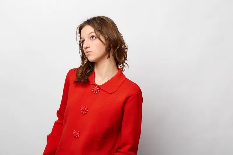 Young woman in red sweater looks away thoughtfully against white background. Casual style, natural light, and minimalistic composition create calm and reflective mood.