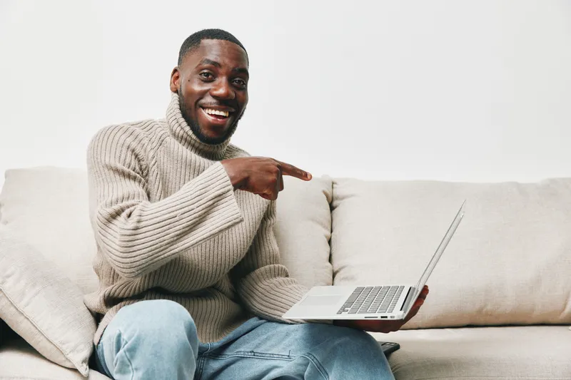 Cheerful Black man in a cozy sweater smiling while pointing at a laptop, representing remote work and modern technology at home