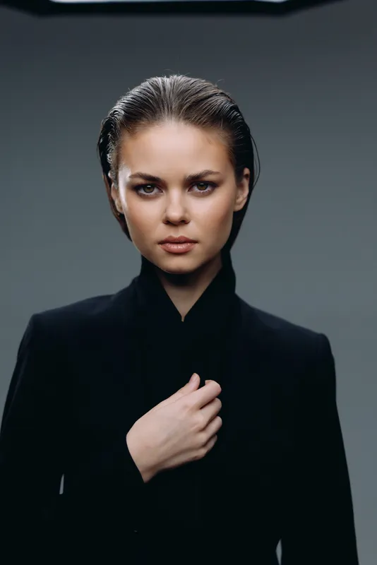 Serious woman in black outfit with slicked-back hair posing with confident expression in studio setting