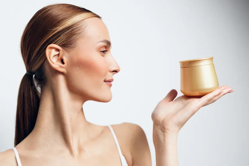 A young woman with clear skin holds a golden cosmetic jar near her face, showcasing skincare and beauty concepts with a clean white background and soft lighting.