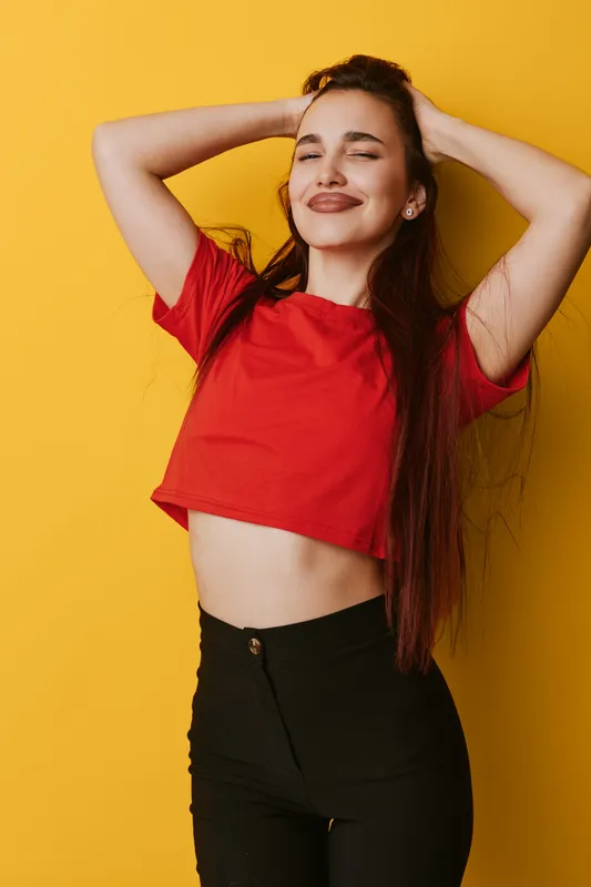 Young woman in red crop top and black pants posing with hands behind head against yellow background. Smiling female model with long hair and closed eyes enjoying moment in studio.