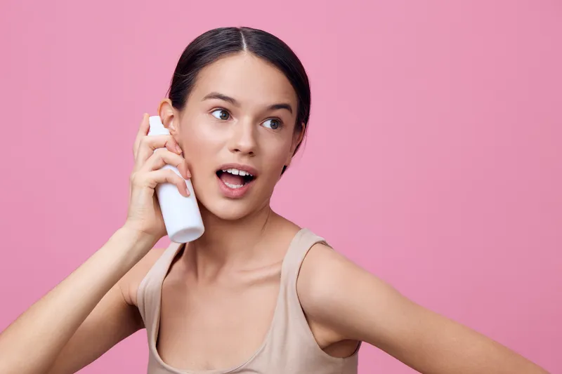 Young female model excitedly talking on the phone with a white spray bottle against a pink background, showcasing trendy fashion and a joyful expression