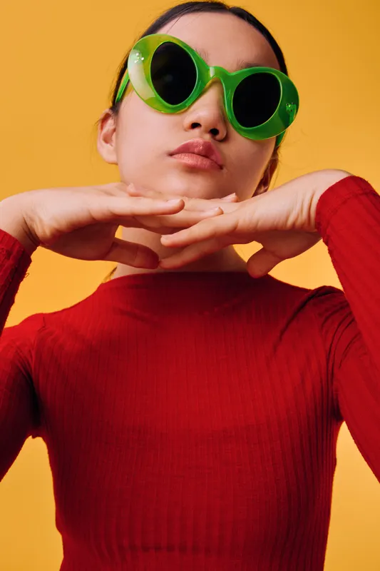 Woman wearing green sunglasses and red sweater posing with hands under chin on bright yellow background. Fashion accessory, style, casual, confident young female portrait with modern outfit.