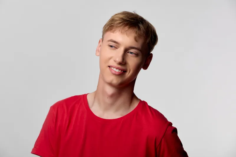 Young man in a red shirt with a thoughtful expression in a studio shot, relaxed pose, and soft lighting delivering a casual modern look.
