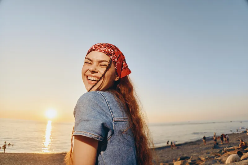 smiling woman in modern boho-western denim jacket enjoys sunset on beach, film color tones, casual lifestyle, natural light, warm mood