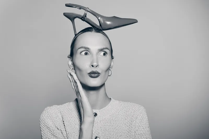 Portrait of a slender white woman with pale skin and slicked hair, wearing a knitted sweater and balancing a pointed high heel shoe on her head. Concept of fashion and quirky style.