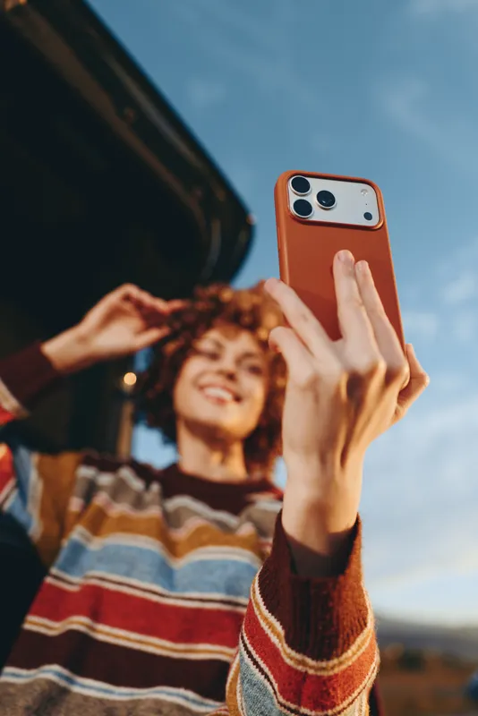 Woman smiles taking a selfie outdoors, holding a smartphone, curly hair and striped sweater, blue sky and warm sunlight in background, lifestyle travel moment with soft focus