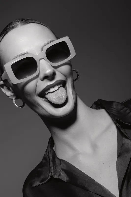 Young white woman with light skin and slicked hair wearing big sunglasses sticks out tongue, smiling in studio. Concept of playful mood and trendy fashion portrait.