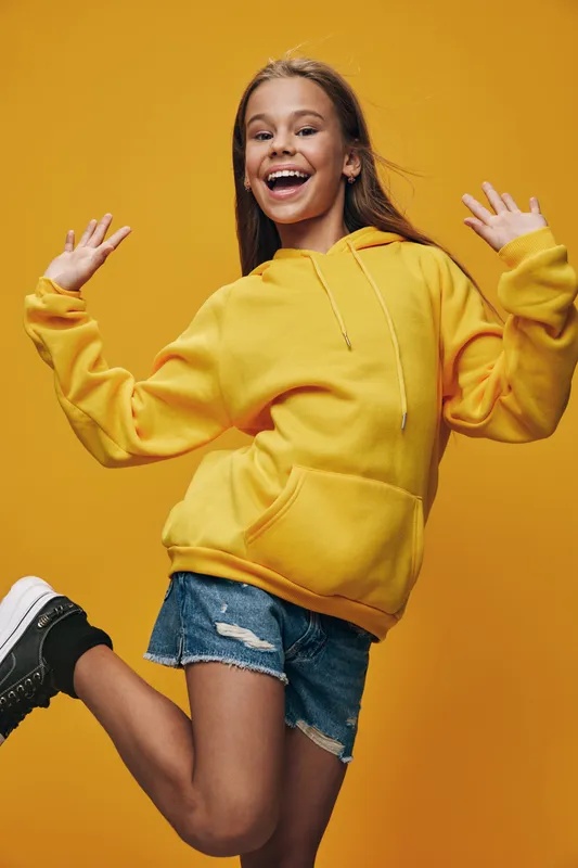 Happy teenage girl wearing yellow hoodie and denim shorts posing energetically on orange background. Smiling young female with casual outfit and joyful expression in studio shot.