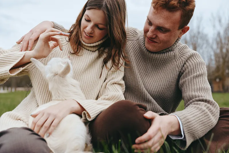 Candid lifestyle shot of a couple in cozy sweaters sitting on grass with a playful goat kid nearby, smiling and gently interacting in a sunny park, capturing warmth and connection together