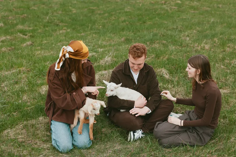 Lifestyle scene with people outdoors, a friendly goat and kid, warm sunlight, relaxed smiles, gentle interaction, grassy field, casual clothing, candid moment, natural colors
