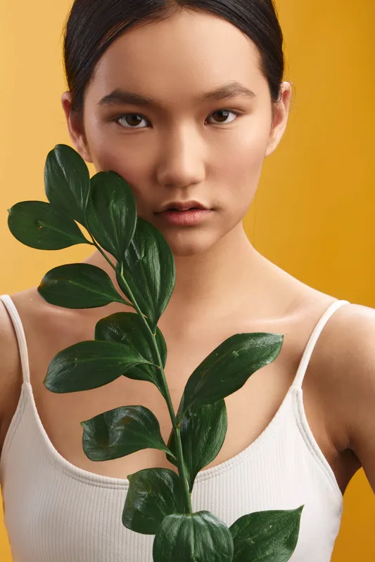 Asian woman holds a leafy branch in front of a warm yellow background, creating a minimal portrait that emphasizes calm poise, natural beauty, and graceful simplicity.