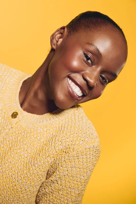 Smiling black woman with dark skin wearing a yellow sweater poses against a bright yellow background. Natural beauty and happiness are emphasized in this vibrant studio portrait.