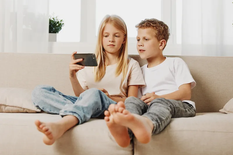 Two children sitting barefoot on a sofa looking at a smartphone together in a bright living room, casual clothing, soft natural light. Concept of family, technology, and childhood.