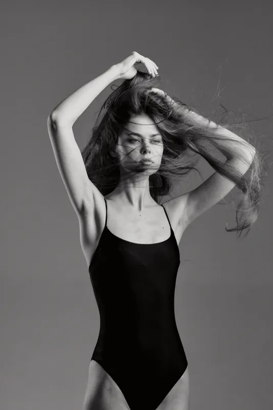 Black and white portrait of a woman in a black swimsuit posing with flowing hair in a minimal studio setting.