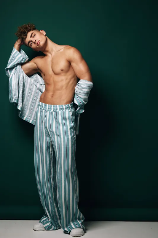 Young man with muscular body posing in striped pajamas shirt open and pants standing against dark background with hand on head and relaxed expression in studio lighting.