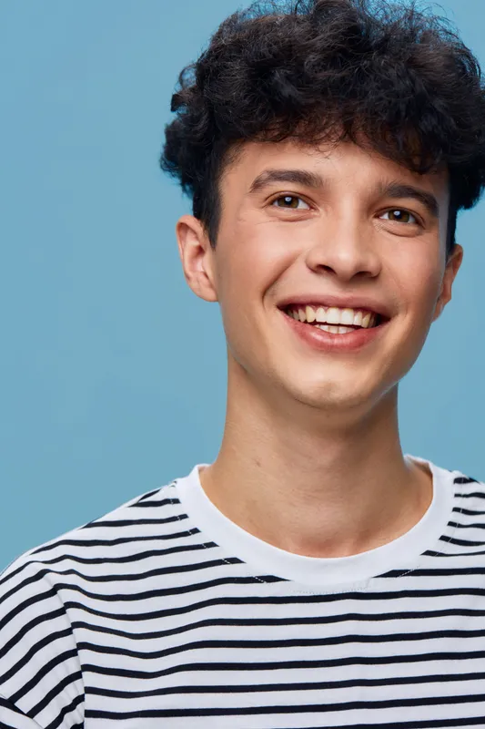 smiling man with curly hair wearing a striped shirt against blue background, cheerful young adult showing happiness and casual style, bright portrait