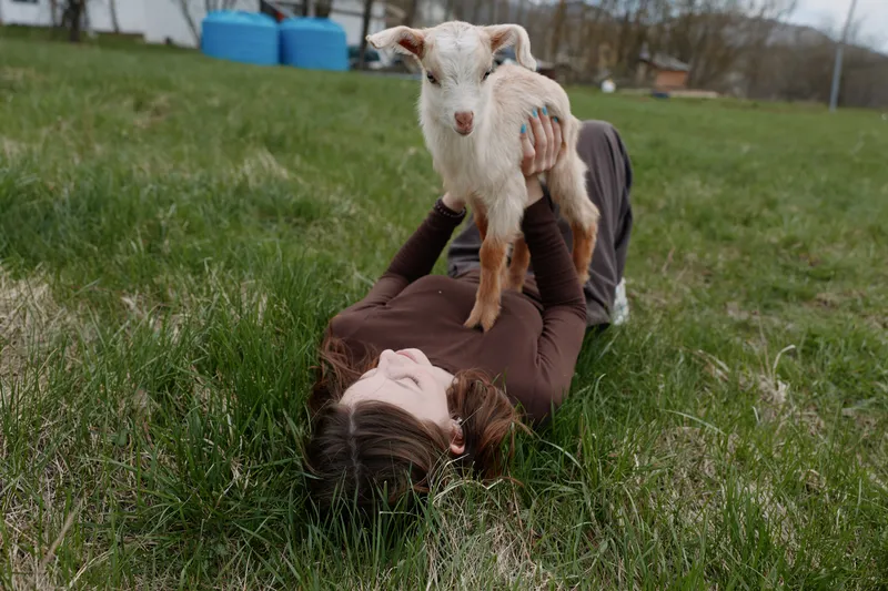 Young woman lies on grass holding a small goat above her with a joyful expression in a natural outdoor setting during a casual lifestyle moment.