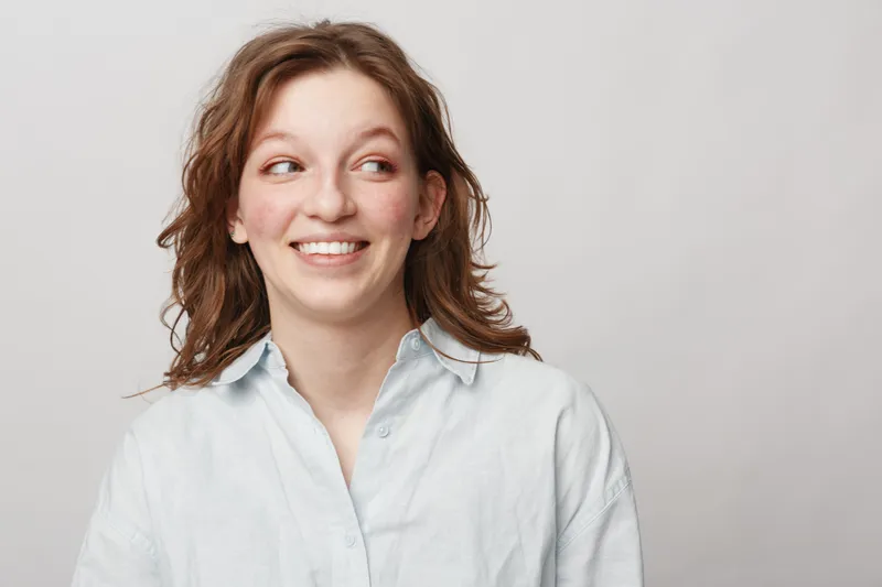 Smiling young woman with curly hair wearing white shirt looks sideways with happy expression on light background. Concept of positive emotions, casual style, and natural beauty.