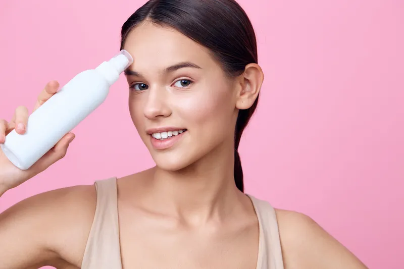 Young woman applying skincare product on her forehead with a smile, against a pink background, showcasing healthy skin and a fresh beauty routine