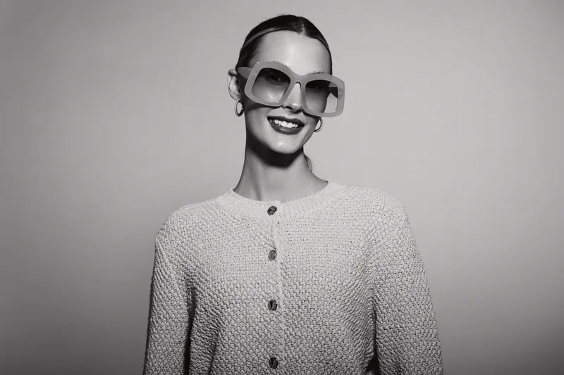 Smiling white woman with fair skin and slicked hair wears large sunglasses and a textured buttoned sweater. Studio portrait on plain background. Concept for fashion and eyewear style.