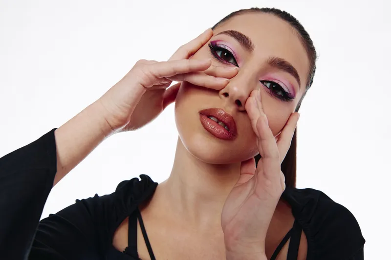 Young woman with artistic pink eye makeup and brown lipstick poses with hands framing her face against a white background. Concept of beauty, creative, and fashion photography.