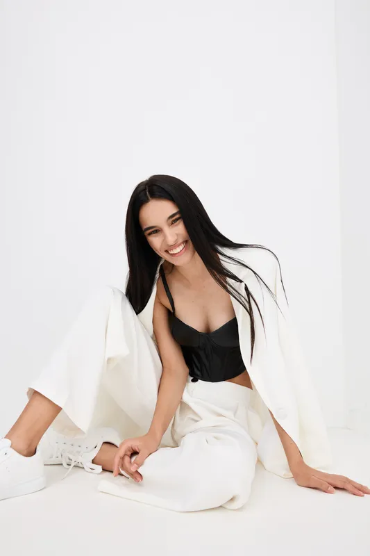 Smiling woman in white outfit sitting on the floor, black top and casual wide-leg pants, cheerful mood and minimalistic bright background, modern fashion style.