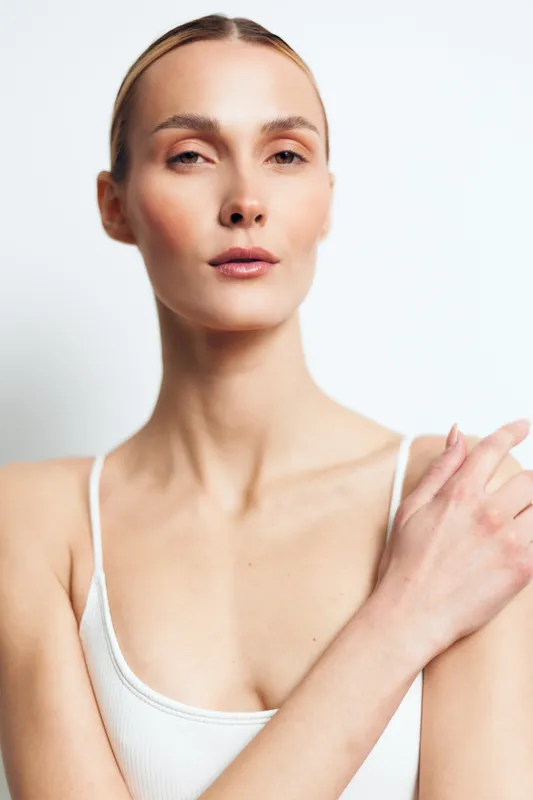 A confident young woman with clear skin and natural makeup looks directly at the camera in a white tank top against a plain background. Concept for beauty and skincare.