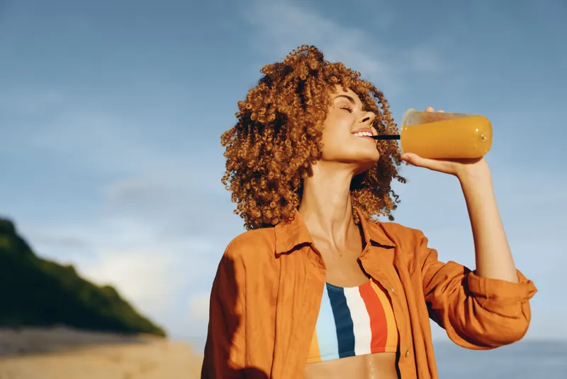 Woman with curly hair drinking juice outdoors by the sea in sunlight relaxed posture warm breeze natural colors casual lifestyle summer travel healthy refreshment