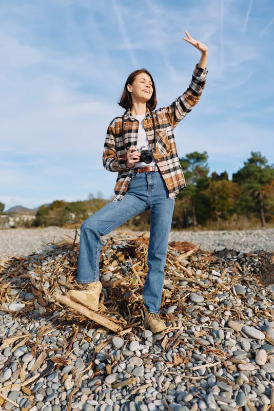 Woman photographer on rocky terrain outdoors, holding a camera and posing with confidence, embracing adventure and nature under a bright sky for creative stock imagery