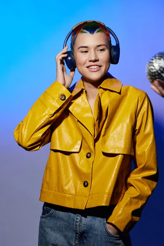 Young woman with colorful hair wearing a yellow jacket listens to music on headphones and holds a disco ball, standing against vibrant blue and purple background, modern style.