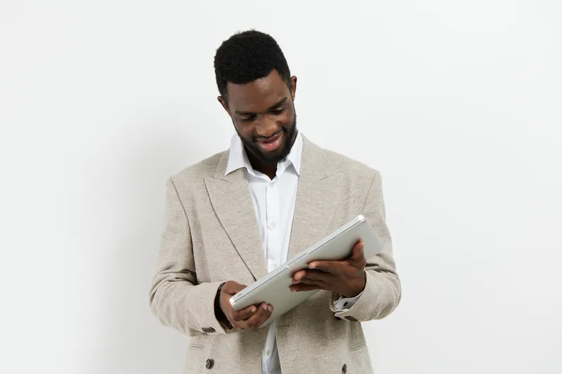 Smiling man with dark skin holding a tablet in hands and looking at screen, dressed in beige blazer and white shirt, standing confidently against plain white background, studio portrait. People lifest