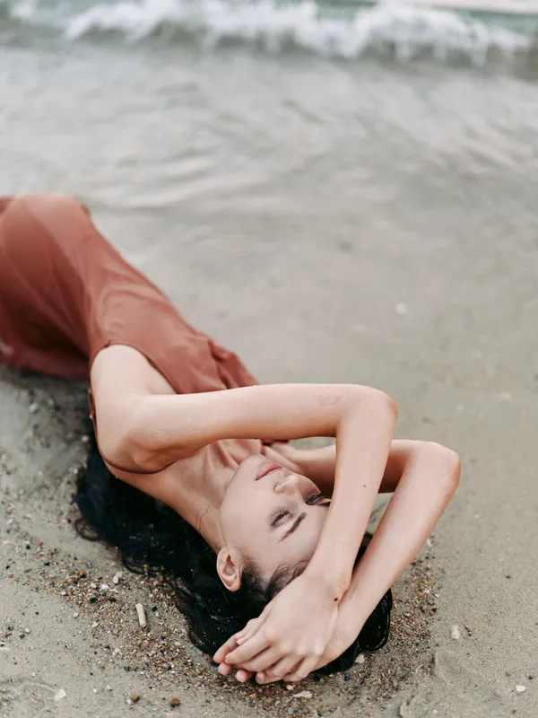 A young woman lies on a sunlit beach, body relaxed and arched along the sand, hair spread, water lapping nearby, creating a serene, intimate moment.