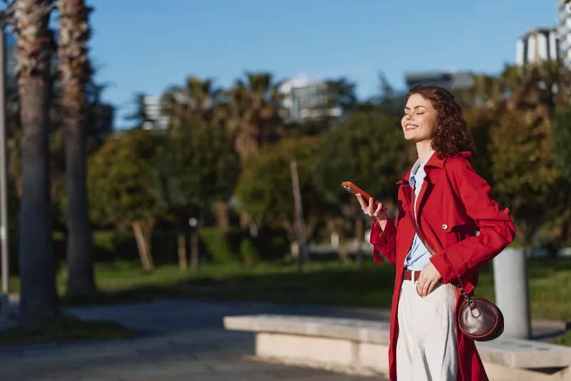 Young woman with curly brown hair in a red coat smiles while holding a phone in a sunlit park, a relaxed casual moment.