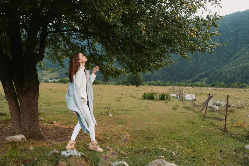 Woman enjoying outdoors under a leafy tree, casual lifestyle moment, breeze in hair, field and mountains in the background, natural serene colors