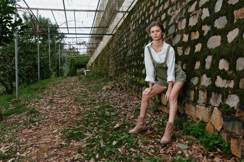 Young woman in casual green overalls and beige boots poses in a serene garden setting, evoking feelings of nature, fashion, and relaxation