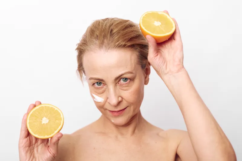 Mature woman with natural skin holds two halves of fresh orange near face with cream on cheek. Concept of healthy lifestyle, skincare routine, natural beauty, and vitamin C benefits.