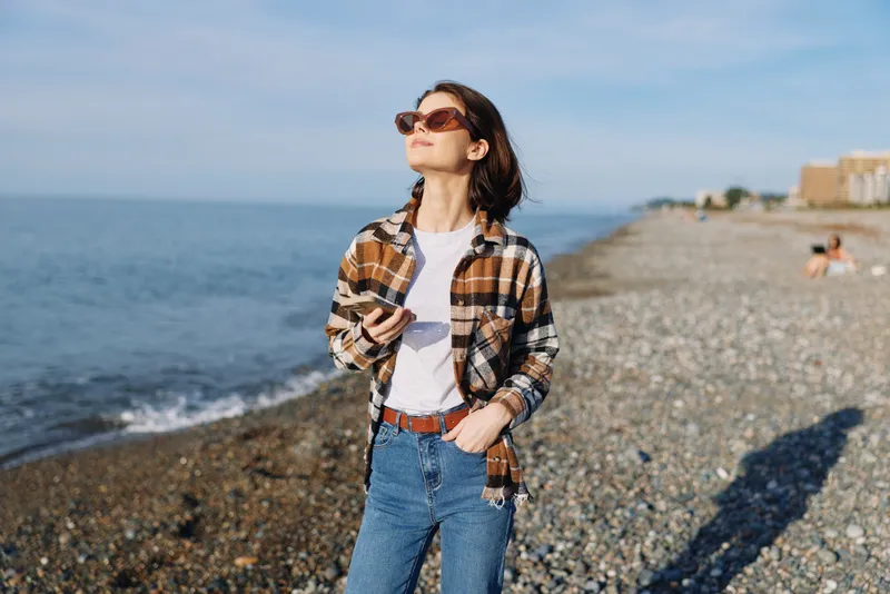Woman in casual plaid shirt and jeans on a pebbled beach, sunglasses on, facing the sun, enjoying the outdoor seaside breeze and blue horizon in a bright portrait style, calm and confident.