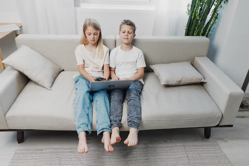 Two children sitting barefoot on a sofa using a laptop in a bright minimalist living room with natural light and casual home atmosphere.